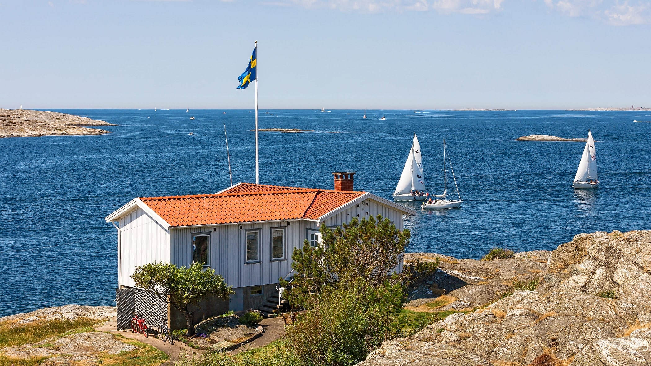 Vit sommarstuga på klipporna vid havet. Svenska flaggan vajar från en flaggstång och på havet seglar två båtar