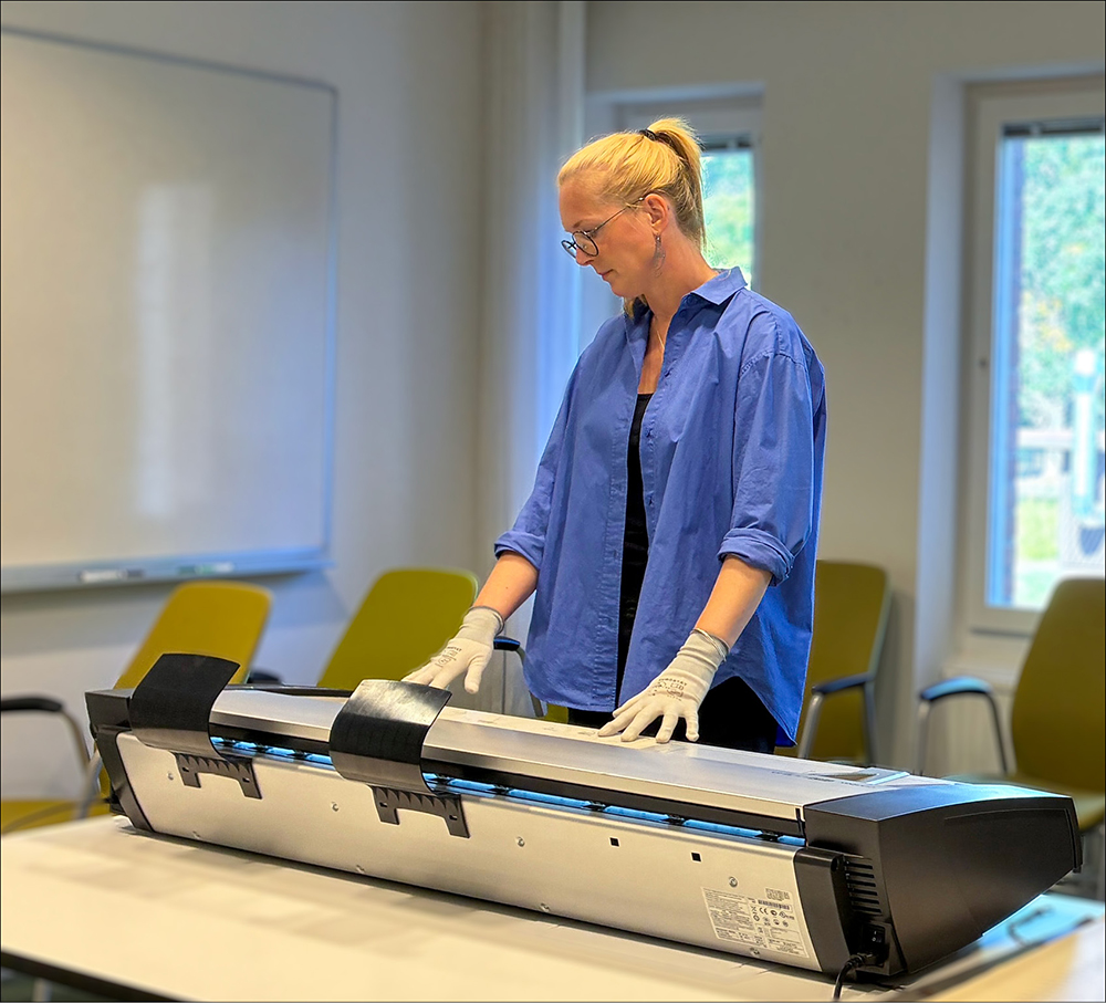 En kvinna med blå skjorta tittar ner på en skrivare som står på ett stort bord.