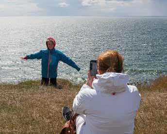 En pojke står på en höjd, men utsikten över havet. En kvinna sitter framför honom och tar en bild med mobiltelefonen.