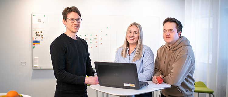 Tre personer, som alla tidigare varit trainees på Lantmäteriet, står i ett mötesrum samlade vid ett litet ståbord och tittar in i kameran. En bärbar dator står uppfälld på bordet. Alla ser nöjda eller glada ut.