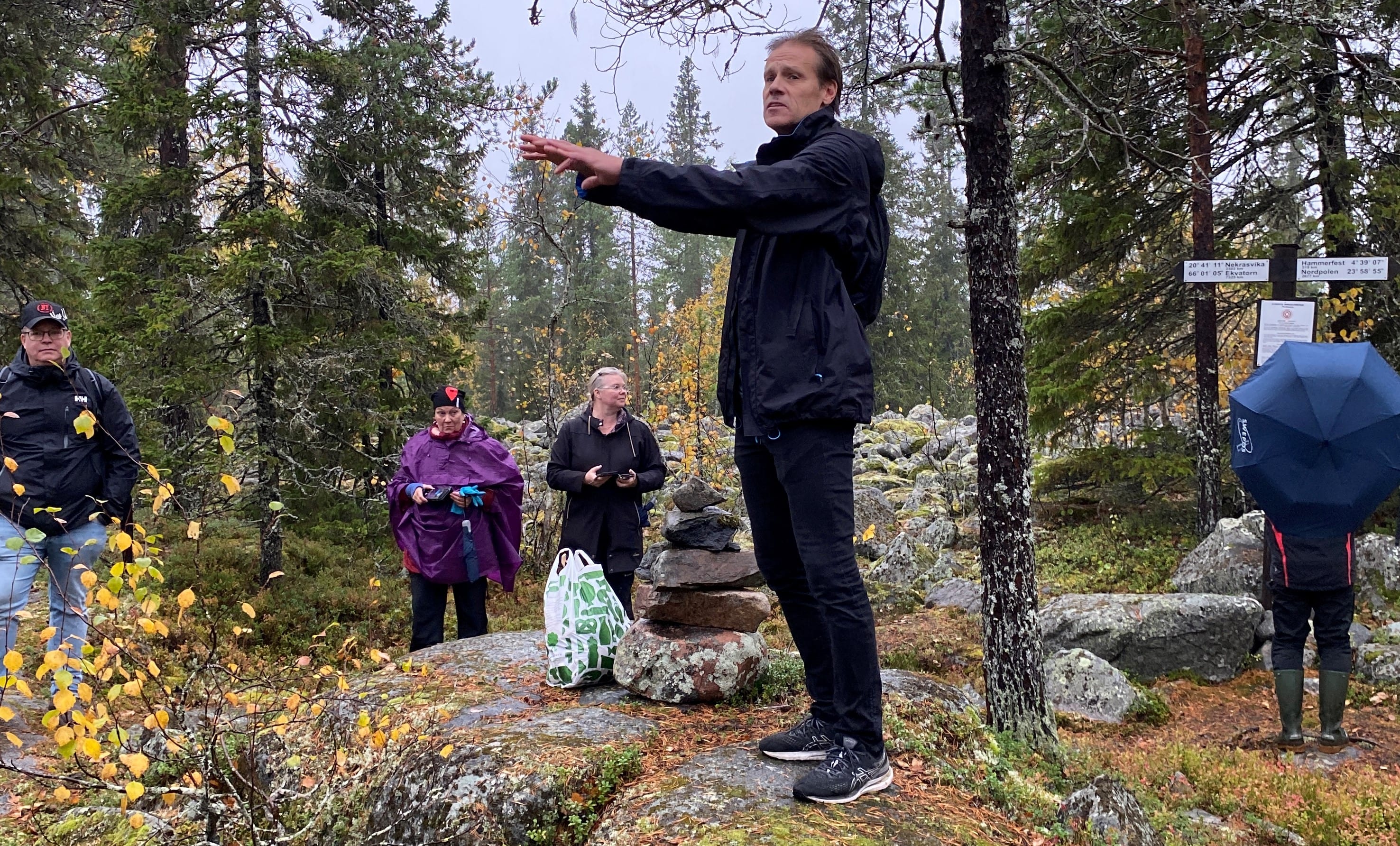En man står vid ett stenröse i tallskogen. Han berättar något för personerna runt omkring.