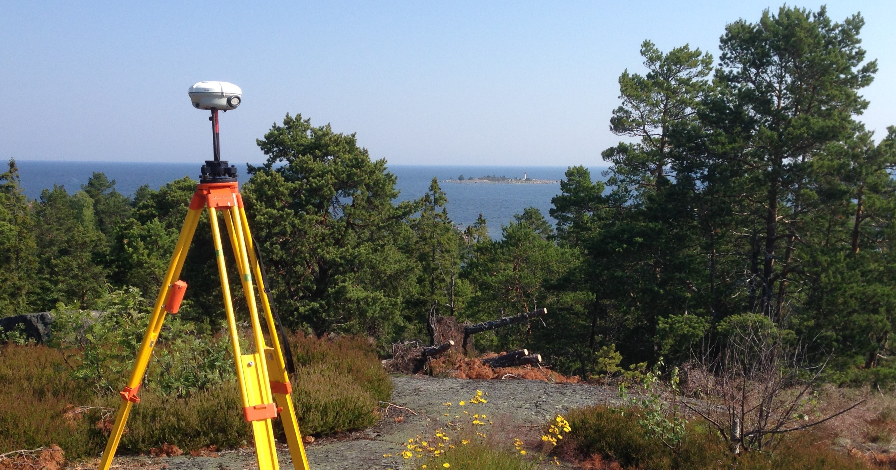 Ett stativ med en GNSS-mottagare står uppställt på en berghäll. I bakgrunden syns några tallar, hav och en liten kobbe.