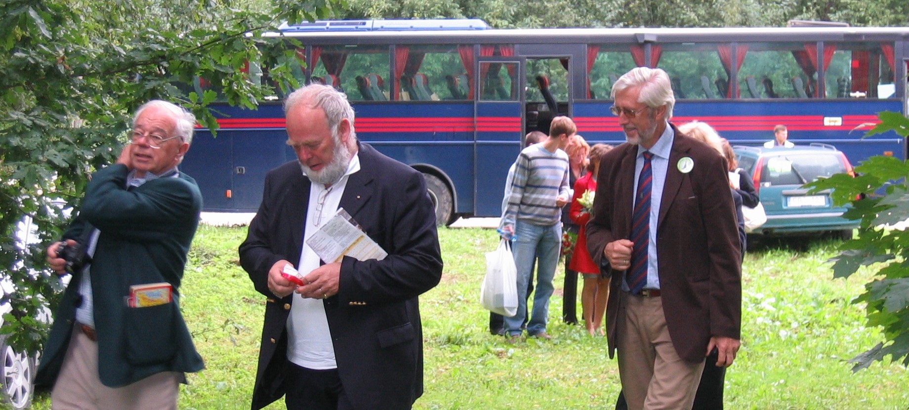 Tre äldre män går över en gräsmatta. I bakgrunden syns en buss och flera människor som står bakom de tre männen.