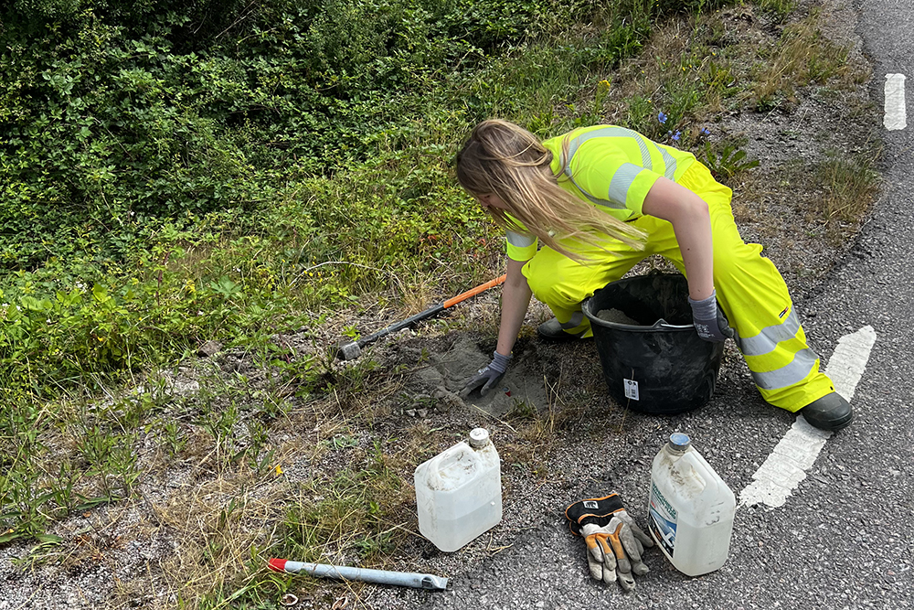 Fair-haired woman wearing work clothes sits by the roadside and collects soil in a bucket. In front of her is a shovel, work gloves and other tools.