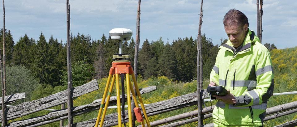 Till vänster står ett stativ med GNSS-antenn monterad. Mätningsingenjören till höger arbetar med fältdatorn och förbereder för statisk GNSS-mätning.