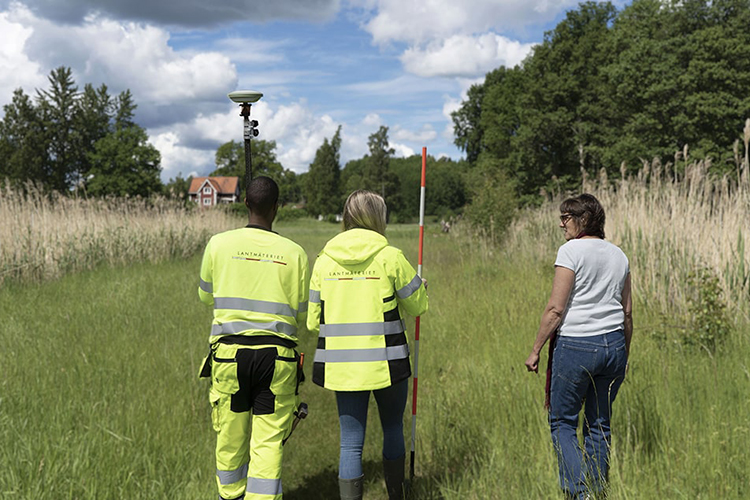 Två lantmätare och en kund står med ryggen mot kameran och tittar ut över en grönskande tomt. Lantmätarna håller mätinstrument i händerna.